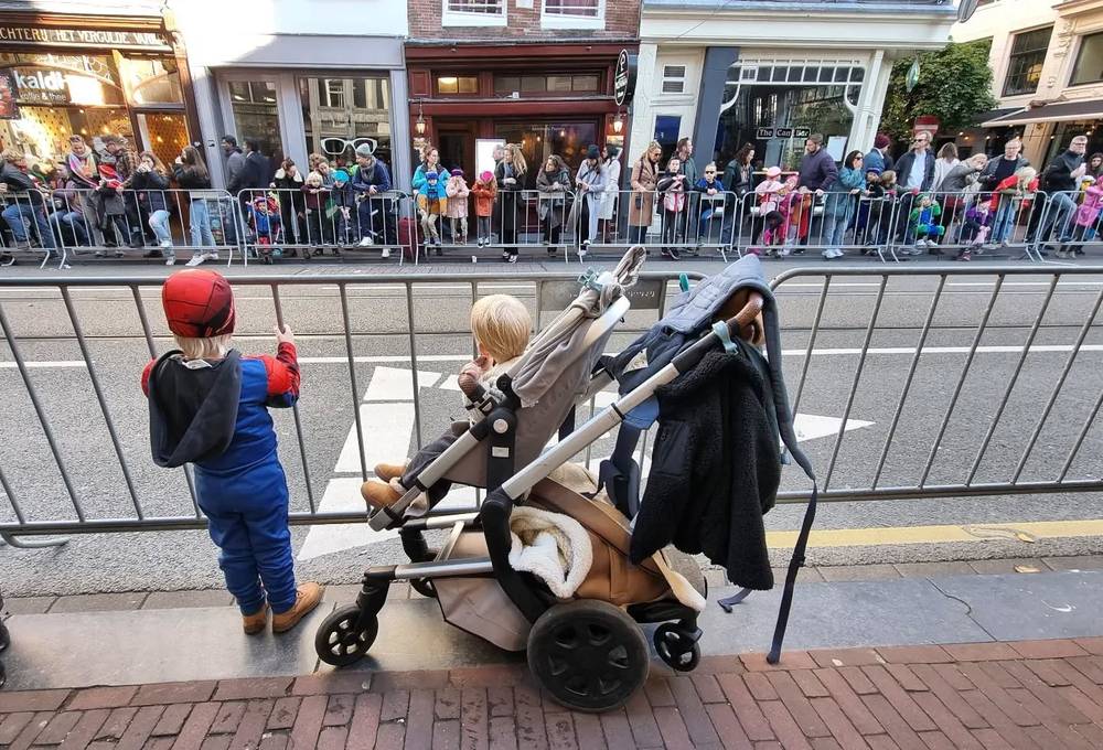 Children in costume watching a street parade from behind a barrier, one dressed as Spider-Man.