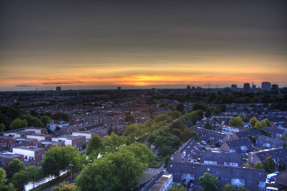 An aerial view over a residential neighborhood with rows of houses and trees, lit by a colorful sunset sky.