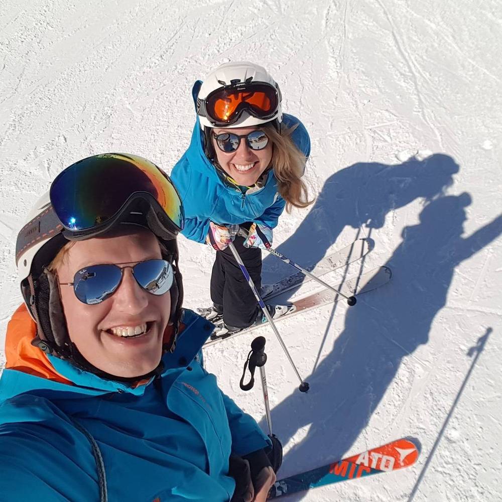 Two skiers smiling for a selfie on a sunny mountain slope.