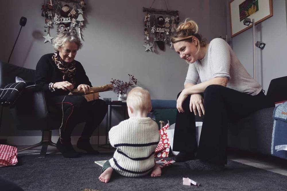 An older woman and a younger woman sit on the floor playing with a baby near Christmas decorations.