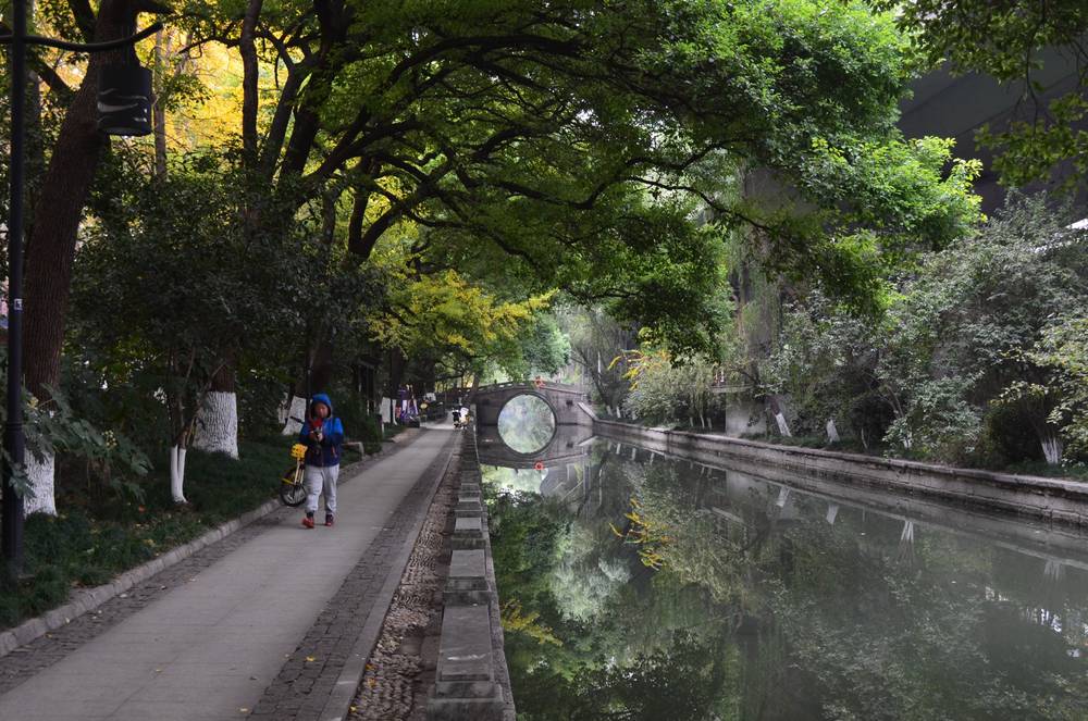 A stone lined canal stretching into the distance, with a canopy of trees reflected perfectly in the still water. A arched bridge forms a perfect circle with its reflection in the distance