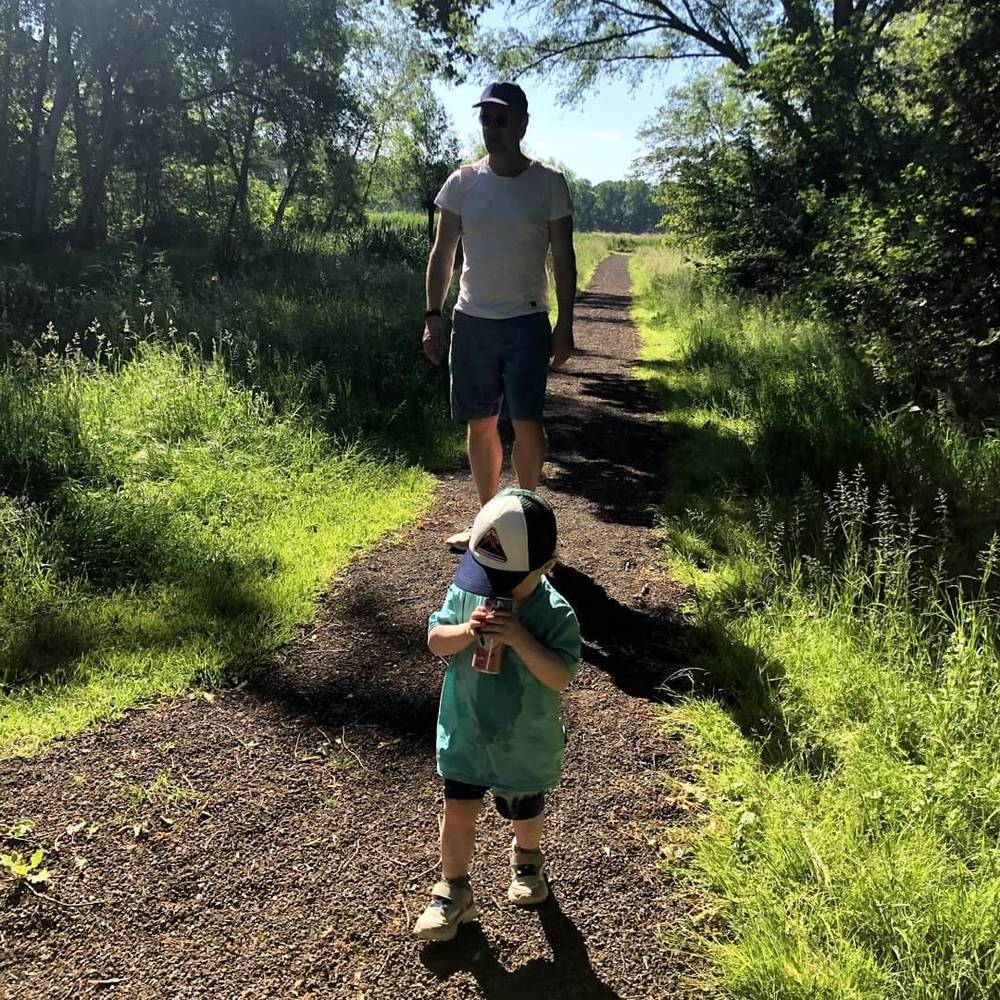 A man walking ahead on a wooded gravel path with a toddler in a cap following behind.