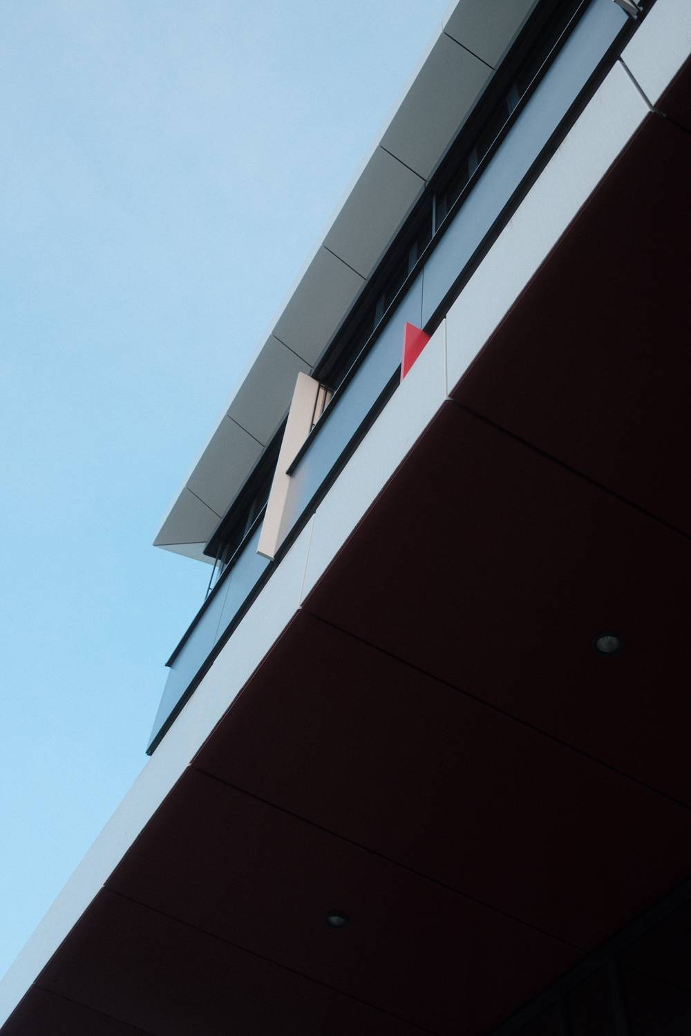 A low-angle view of a modern building's exterior corner against a pale blue sky, featuring dark red cladding and a small red triangular sign.