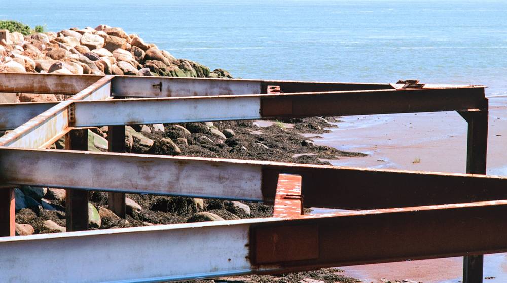 Rusted steel beams of an old pier frame a view of seaweed-covered rocks, wet sand, and the bright blue sea.