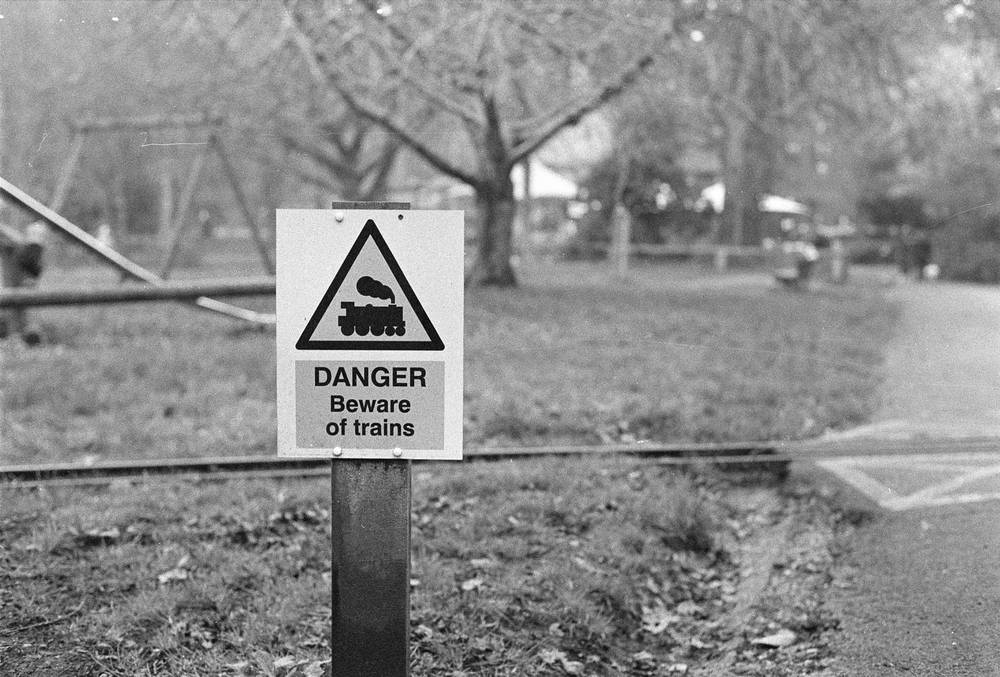 A black and white photograph of a small sign that says "DANGER - Beware of trains". Narrow-gauge railway tracks are visible running across the frame behind the sign.