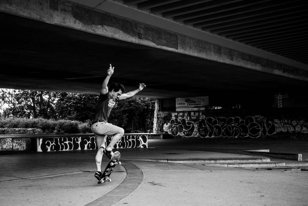 Black and white photo of a skateboarder in mid-motion balancing dynamic pose on a skateboard with arms raised, set in an urban skatepark under a wide concrete bridge covered in graffiti.