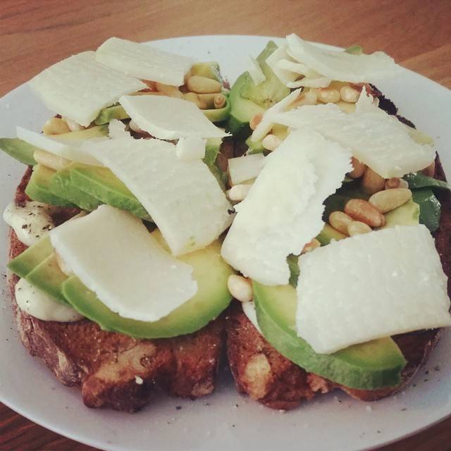 Toasted bread topped with avocado, shaved cheese and pine nuts on a plate.