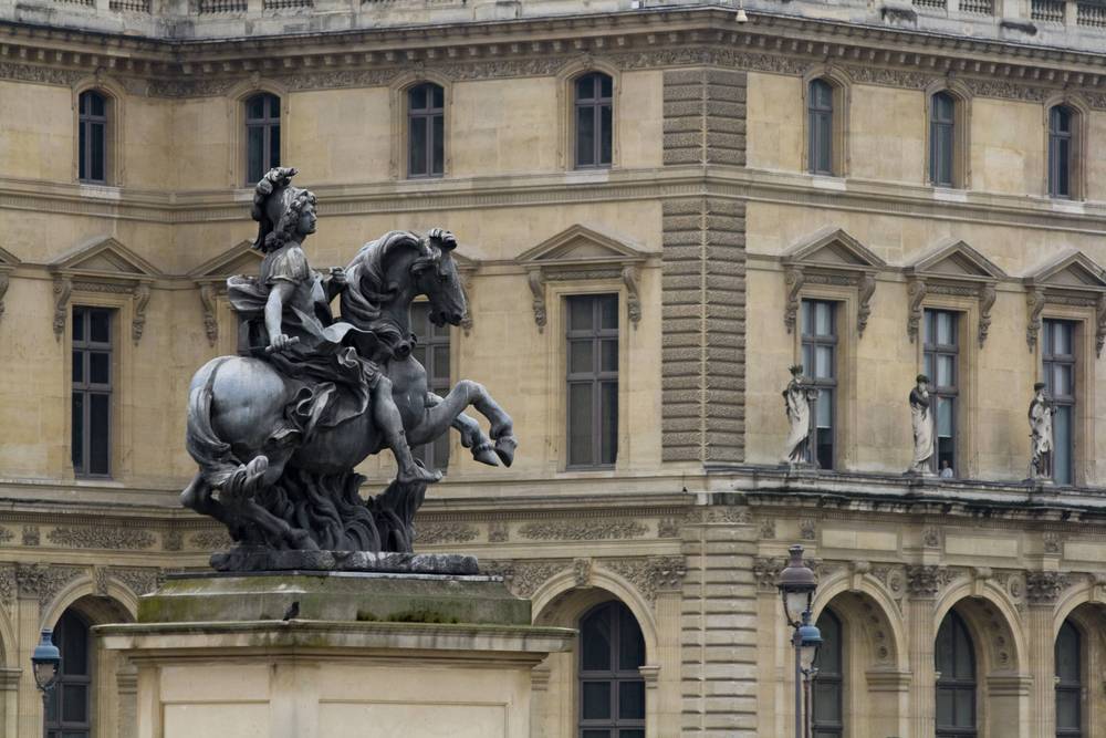 A bronze equestrian statue of a king stands atop a pedestal in front of the Louvre's facade.