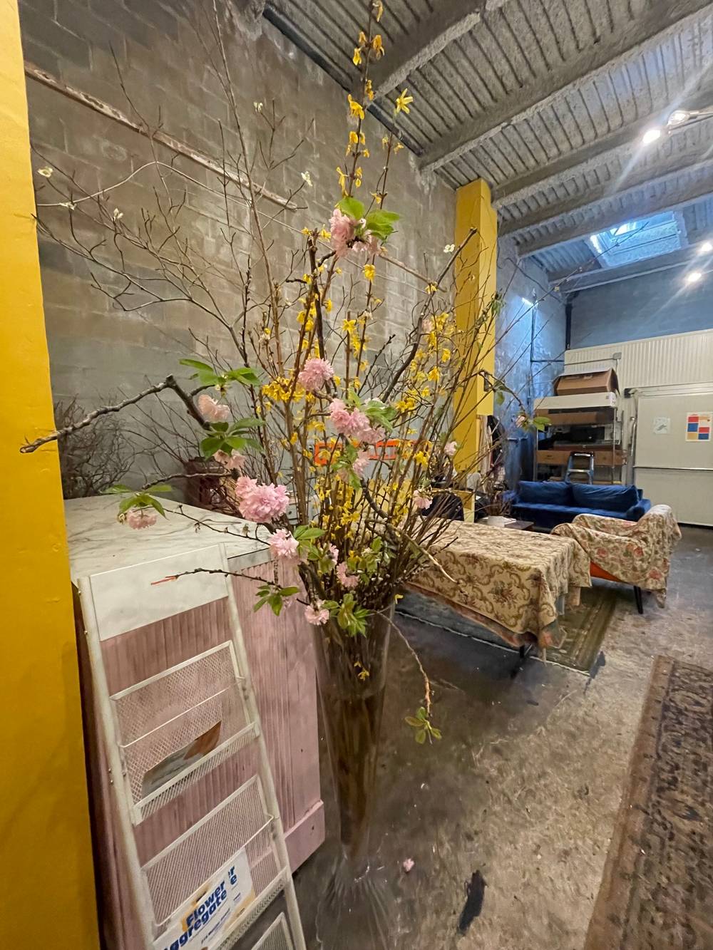 inside a warehouse with concrete walls. a large vase with yellow and pink flowers that are over 10 feet