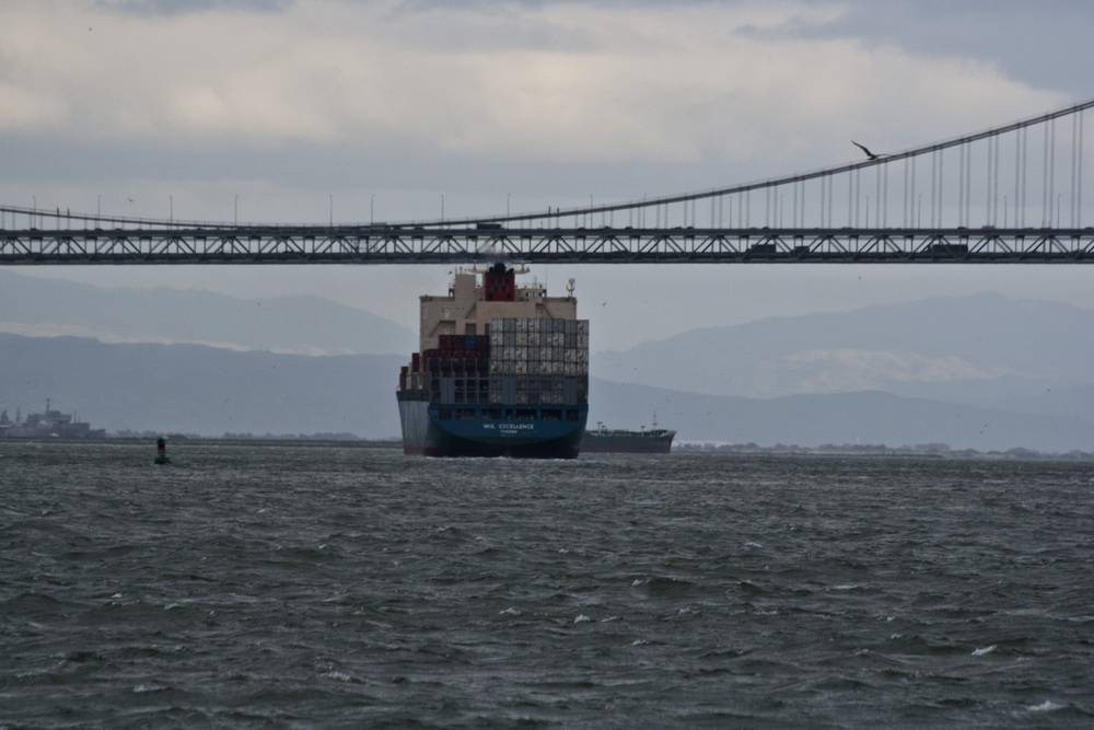 A container ship passes beneath the Bay Bridge on choppy water.