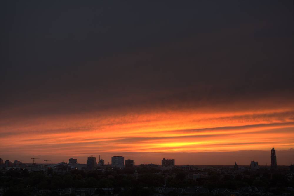 An orange sunset glow behind the dark silhouette of the Utrecht skyline, seen from a rooftop.