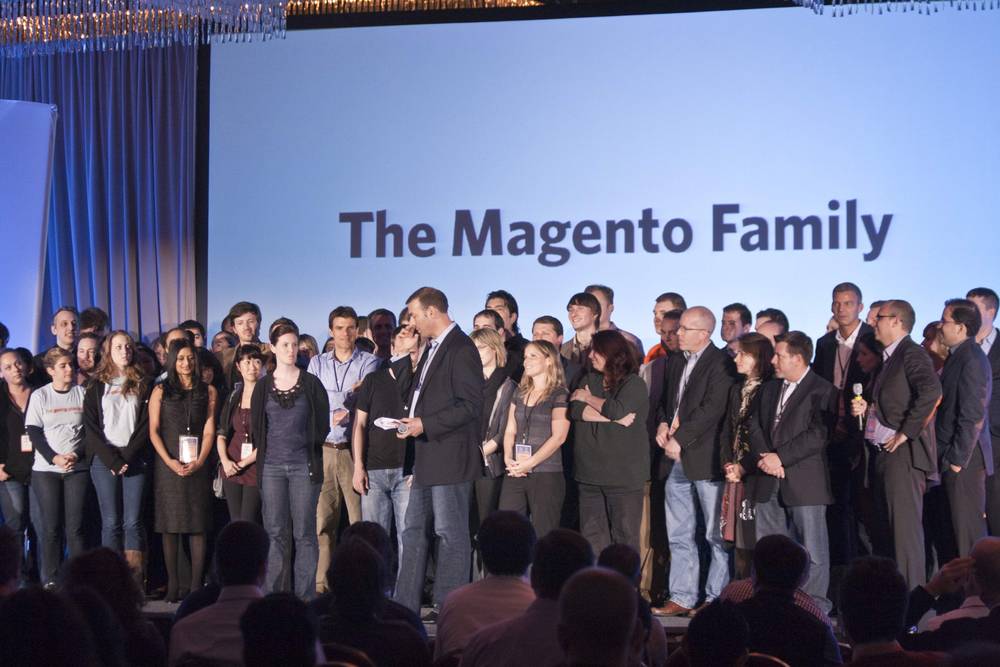 A large group of Magento staff stands on stage beneath a screen reading 'The Magento Family.'