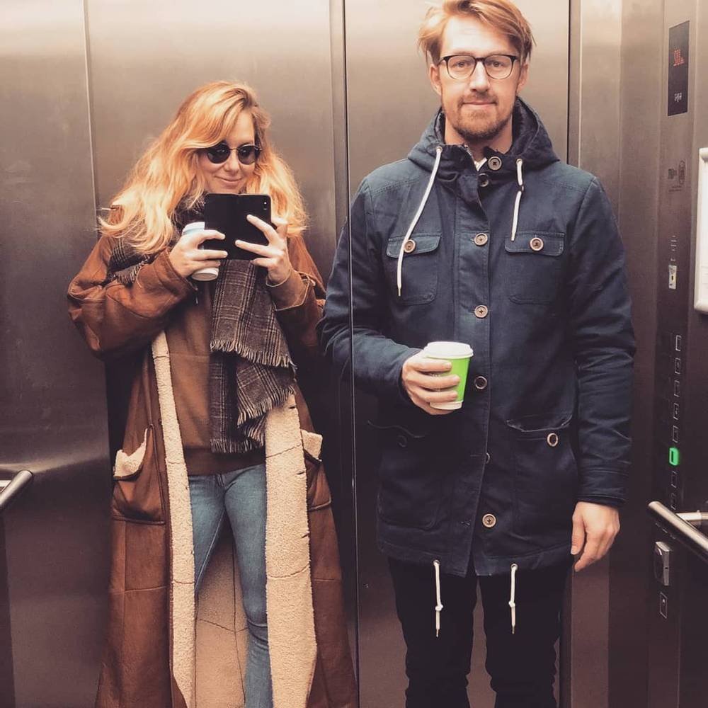 A couple takes a mirror selfie in an elevator, both holding coffee cups.