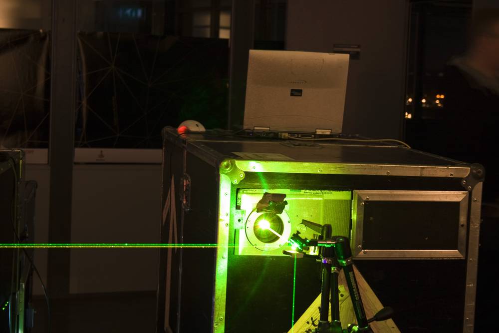 A laser projector rig with a green beam sits on a rooftop workspace, laptop on top.