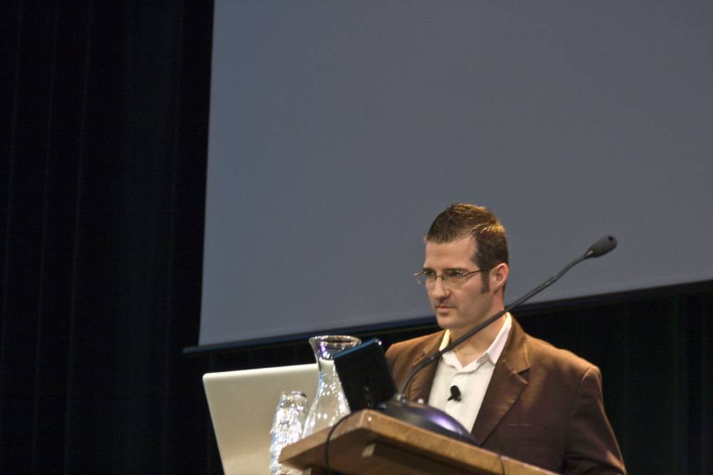 A speaker in a brown jacket addresses the audience at a lectern.