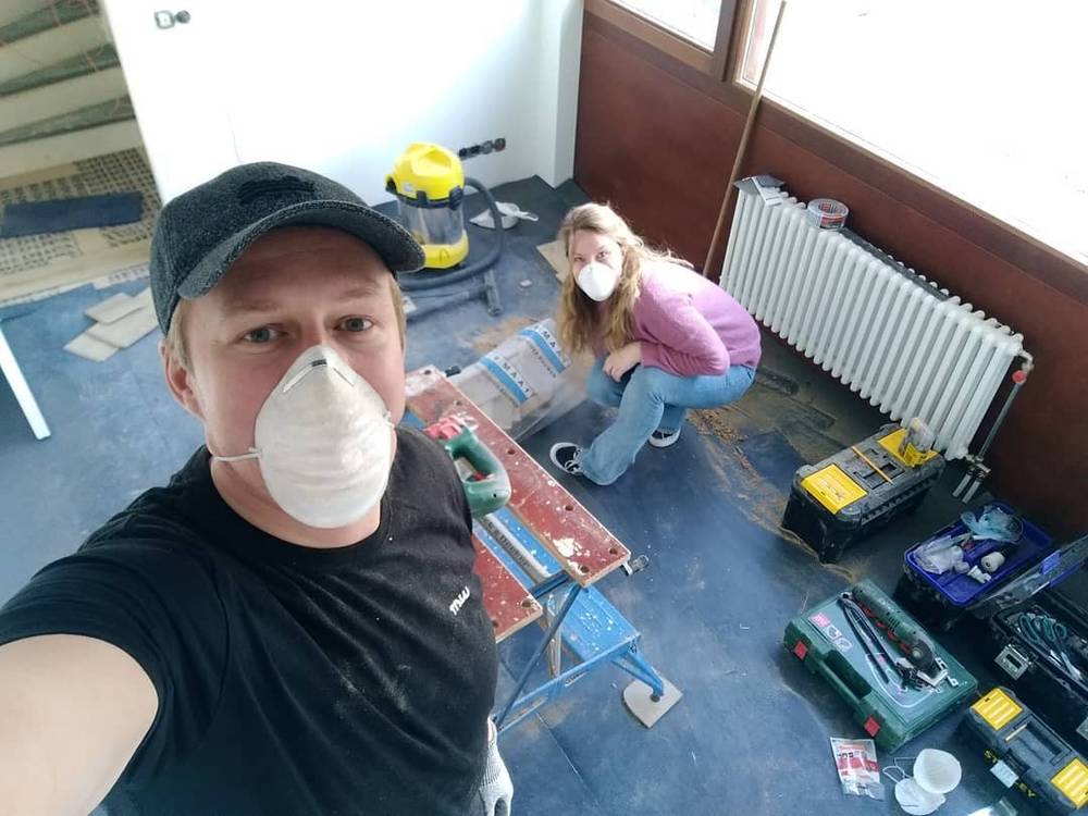 Selfie wearing a dust mask during a home renovation, with a woman working in the background.