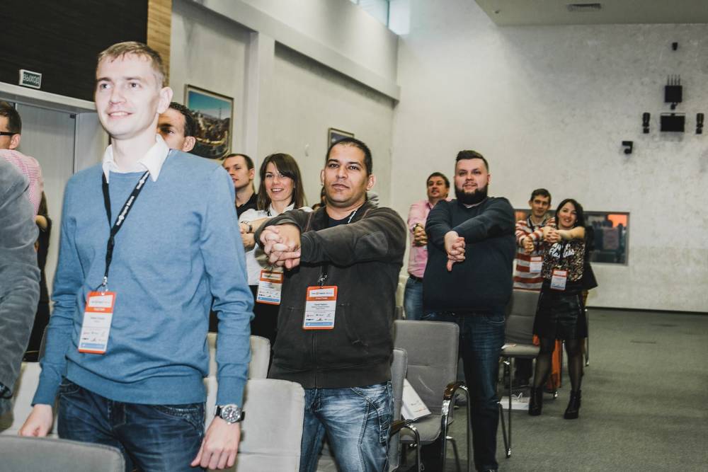 Conference attendees clasp hands together while standing among rows of chairs.