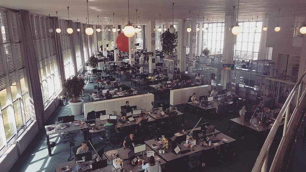 A wide view of a modern open-plan office with rows of desks, hanging lights, and large windows.