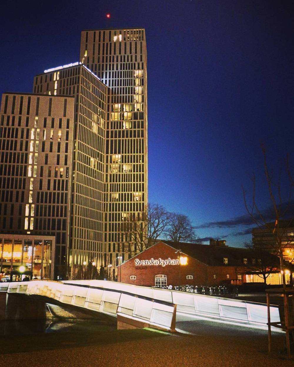 A modern illuminated high-rise building at dusk, reflected in water.