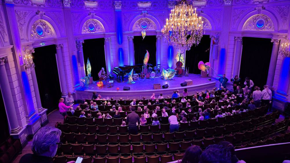 An ornate concert hall interior with a large audience and chandeliers, seen from a balcony.