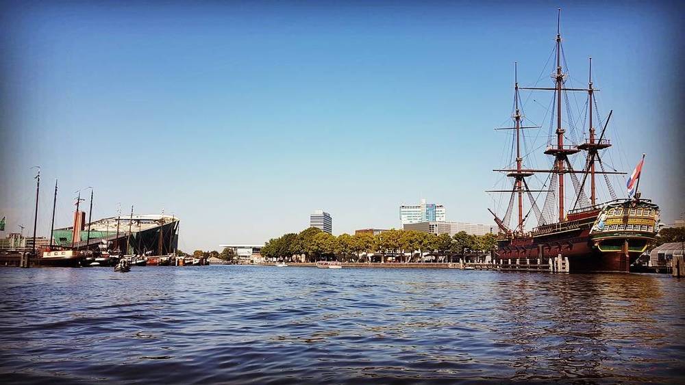 A tall sailing ship moored along an Amsterdam waterway with the city skyline behind it.