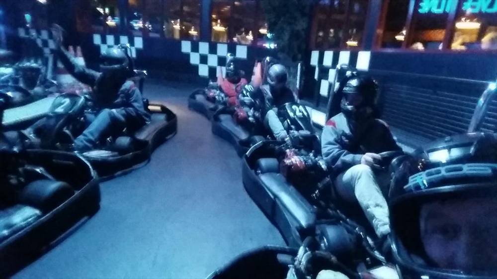 An indoor go-kart track with racers in helmets driving past checkered flag decor.