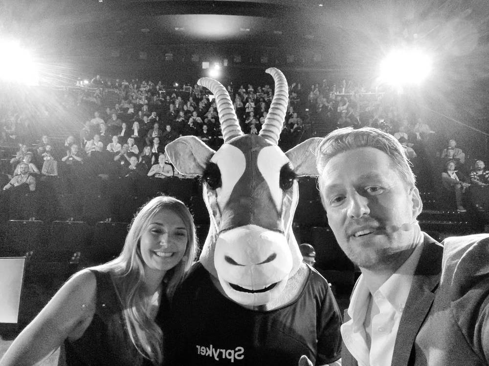 A black-and-white selfie with a woman and someone in a goat mascot costume, with a cheering crowd behind them.