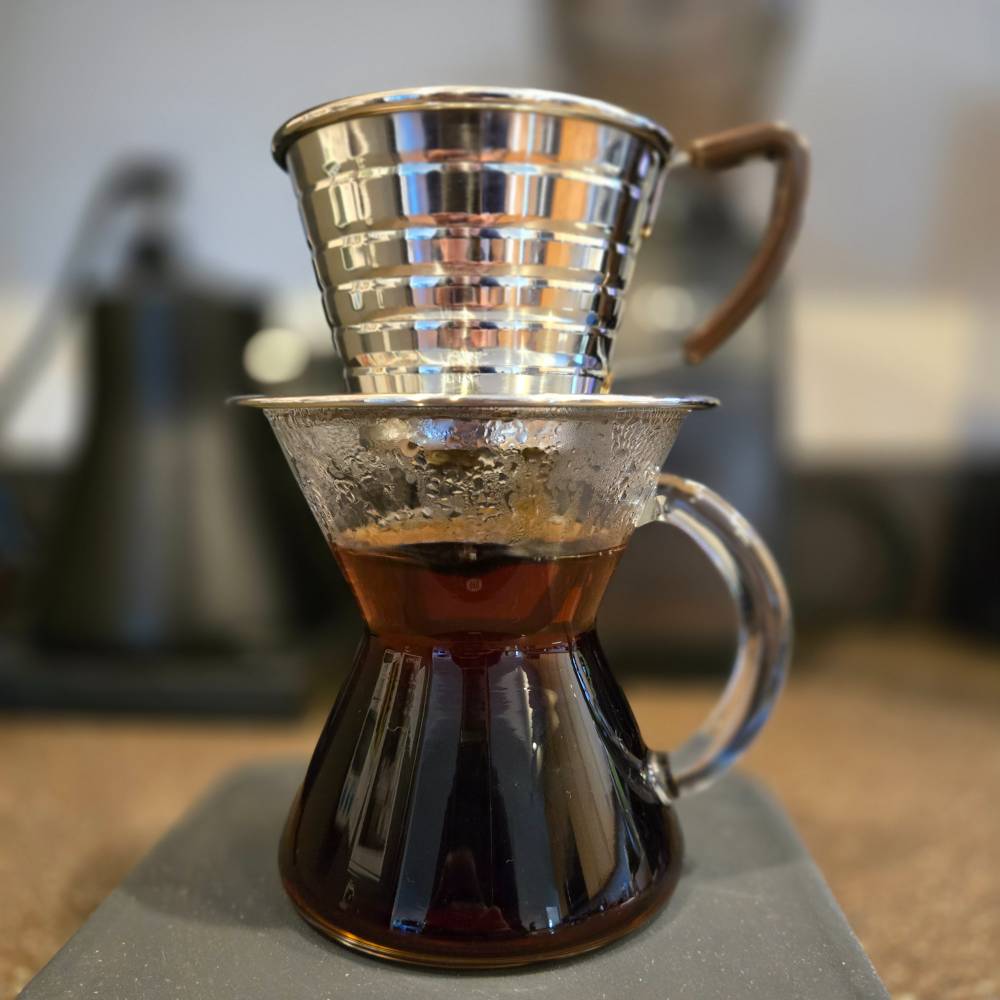 A pour-over coffee setup with a polished stainless steel dripper resting on a clear hourglass-shaped glass carafe, nearly full with dark brewed coffee. A blurred kettle is visible in the background.