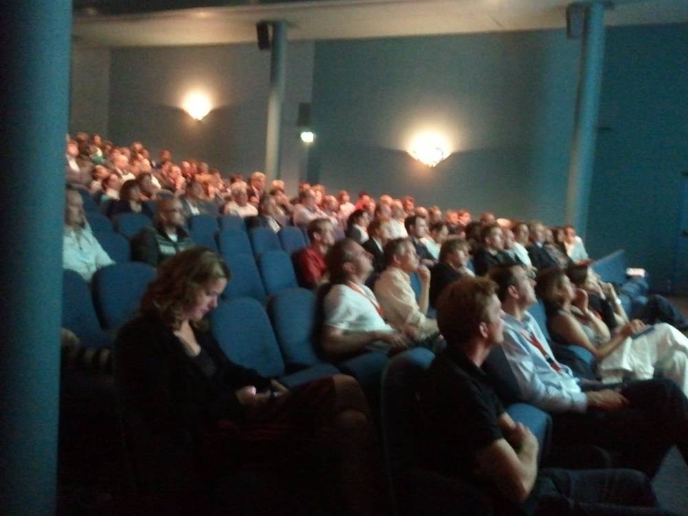 A packed theater audience watches a presentation from blue seats.