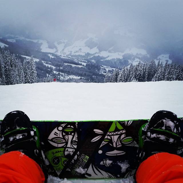 View over snowboard bindings looking out across a snowy valley with forested slopes.