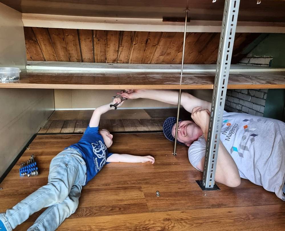 Two people lying on their backs on a wooden floor, reaching up to work on shelving in an attic.