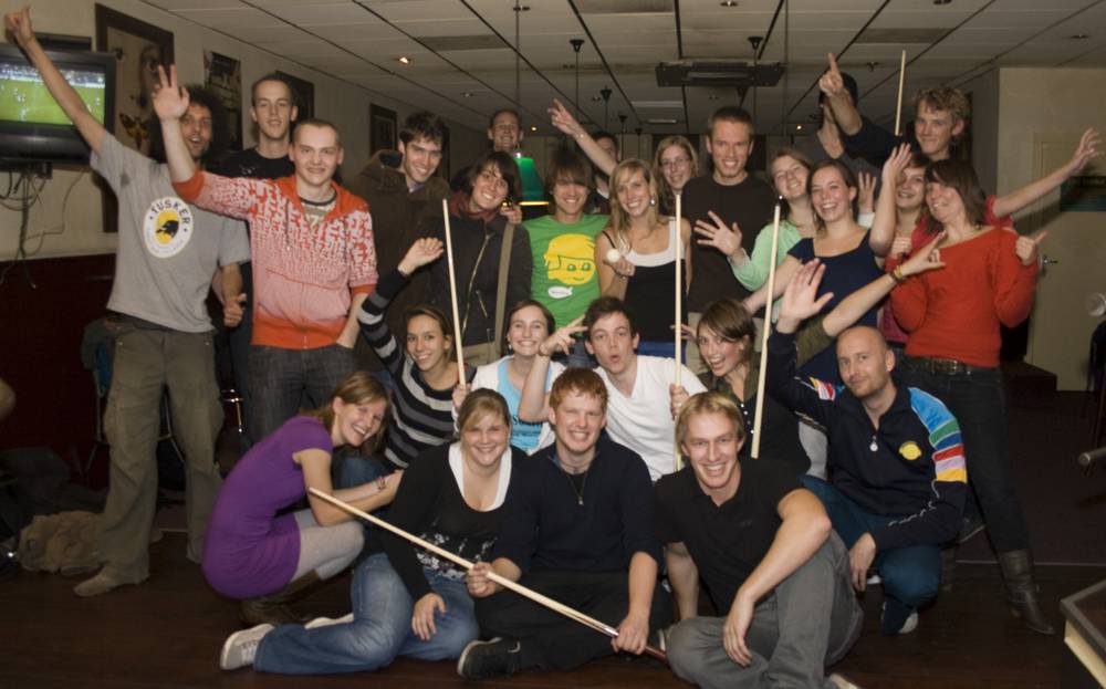 A large group photo of friends posing with pool cues in a bar, arms raised in celebration.