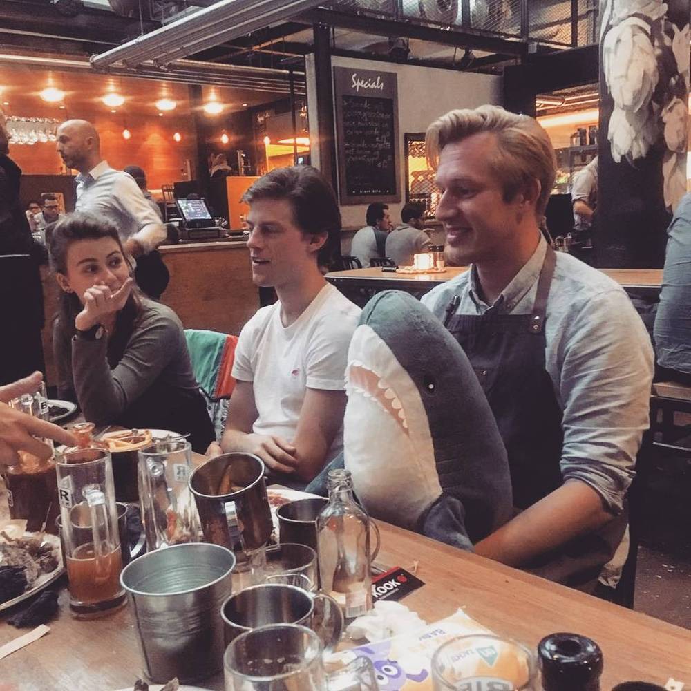 Friends laughing together at a restaurant table, with empty glasses and a shark-shaped cushion.