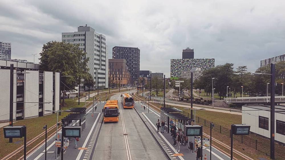 A city street with orange trams passing between modern buildings under a cloudy sky.