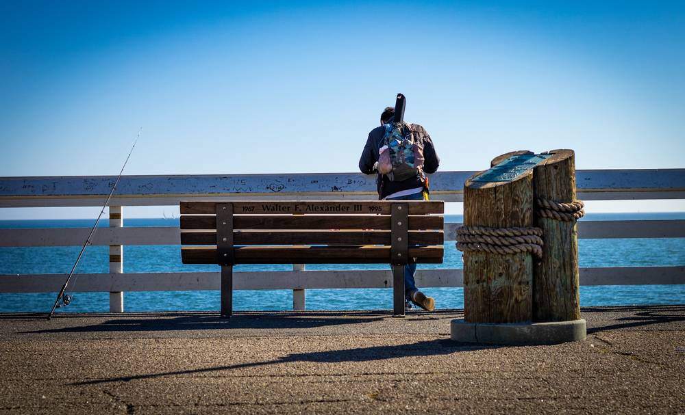 A person carrying a backpack is standing in front of a memorial bench on a pier dedicated to “Walter F. Alexander III, 1967–1995.” A fishing rod leans against the railing to the left, and rope-wrapped wooden pilings stand to the right, with calm blue ocean and clear sky in the background.