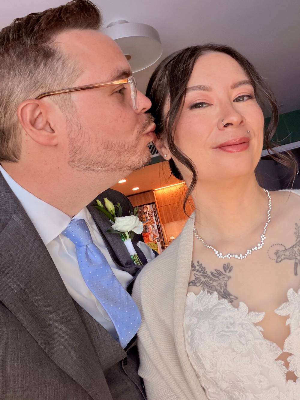 Close-up selfie of a groom in a gray suit and blue polka-dot tie kissing his bride on the cheek — she wears a lace wedding dress, a diamond necklace, and has floral chest tattoos, and accepts the kiss with a satisfied smile