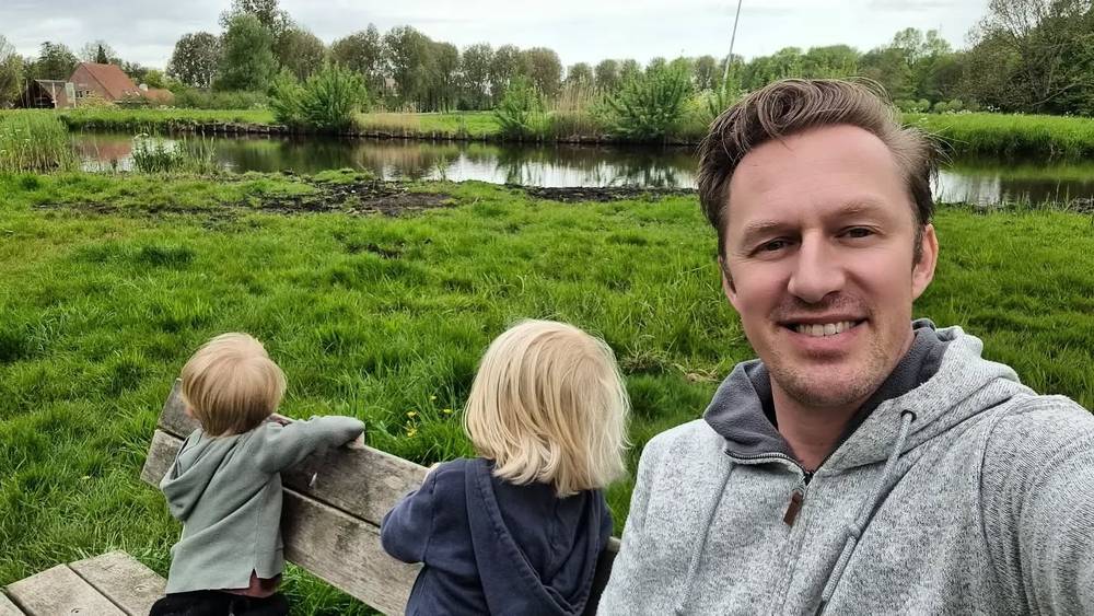 Guido smiling in a selfie with two children looking over a fence at a grassy field and pond.