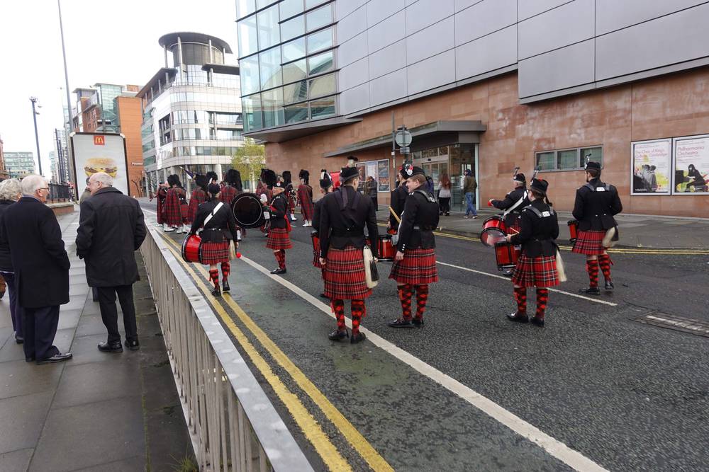 Bagpipers and drummers in ceremonial dress march past onlookers in Manchester.