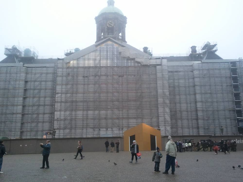 Amsterdam's Royal Palace on Dam Square stands wrapped in scaffolding for renovation, with visitors below.