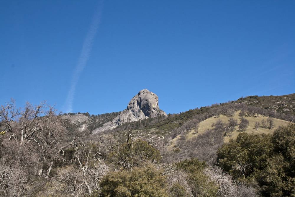A large granite dome rises above a hillside of bare trees and scrub.