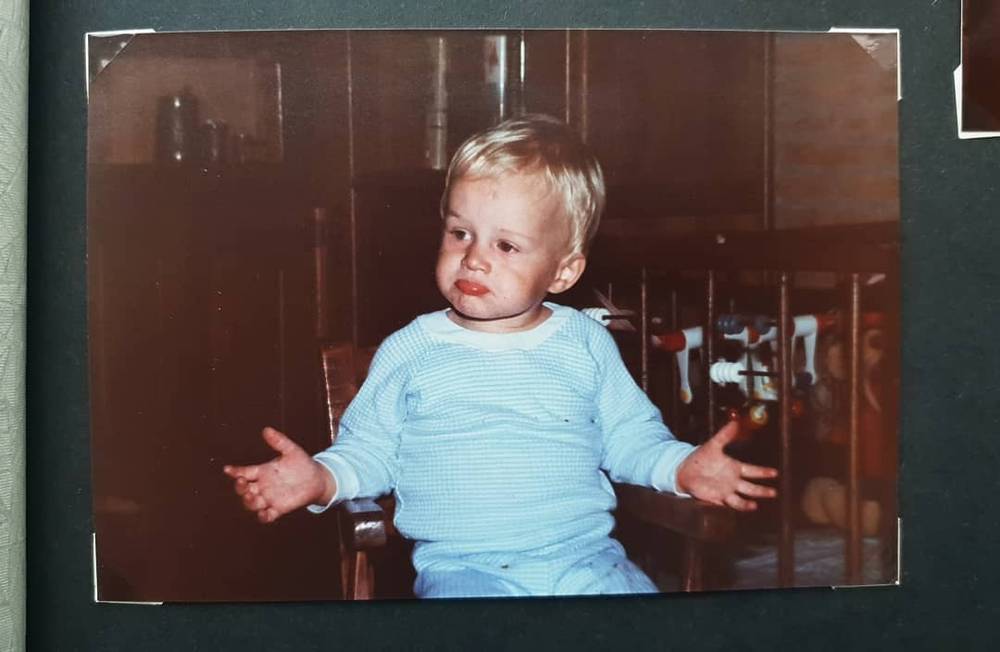 An old photo of a toddler in a light blue sweater sitting with arms outstretched.