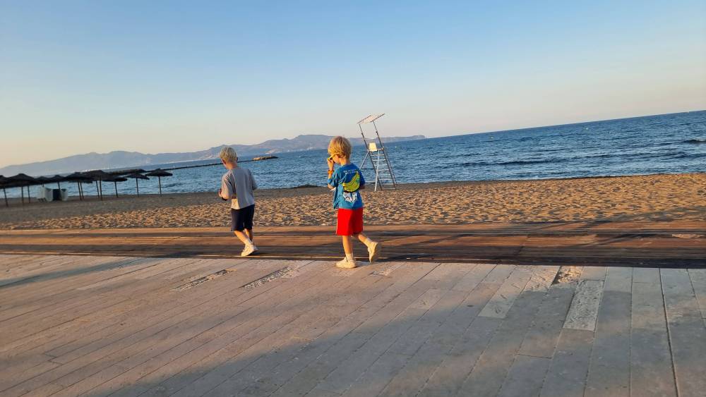 Two children walk along a wooden boardwalk beside a beach at sunset.