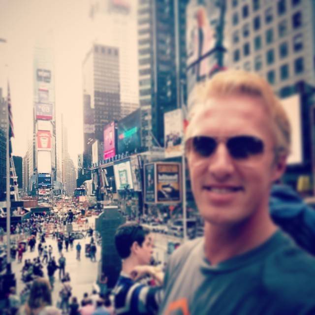 A smiling man in sunglasses poses in front of a crowded Times Square in New York City.