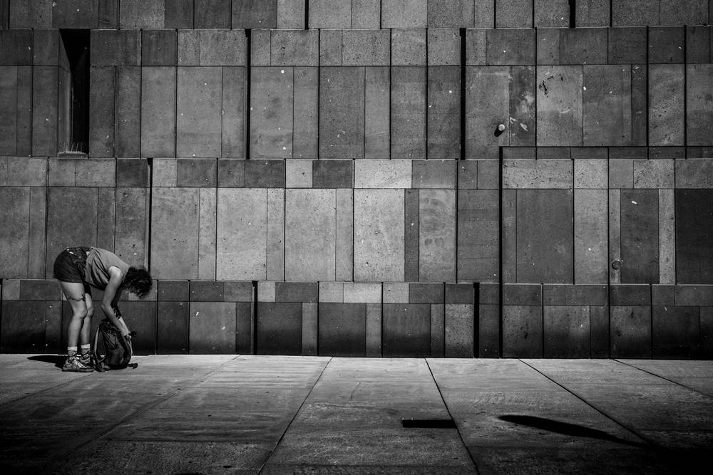 A high-contrast black and white street photograph of a person bending down to reach into a backpack on the pavement against a large stone wall.