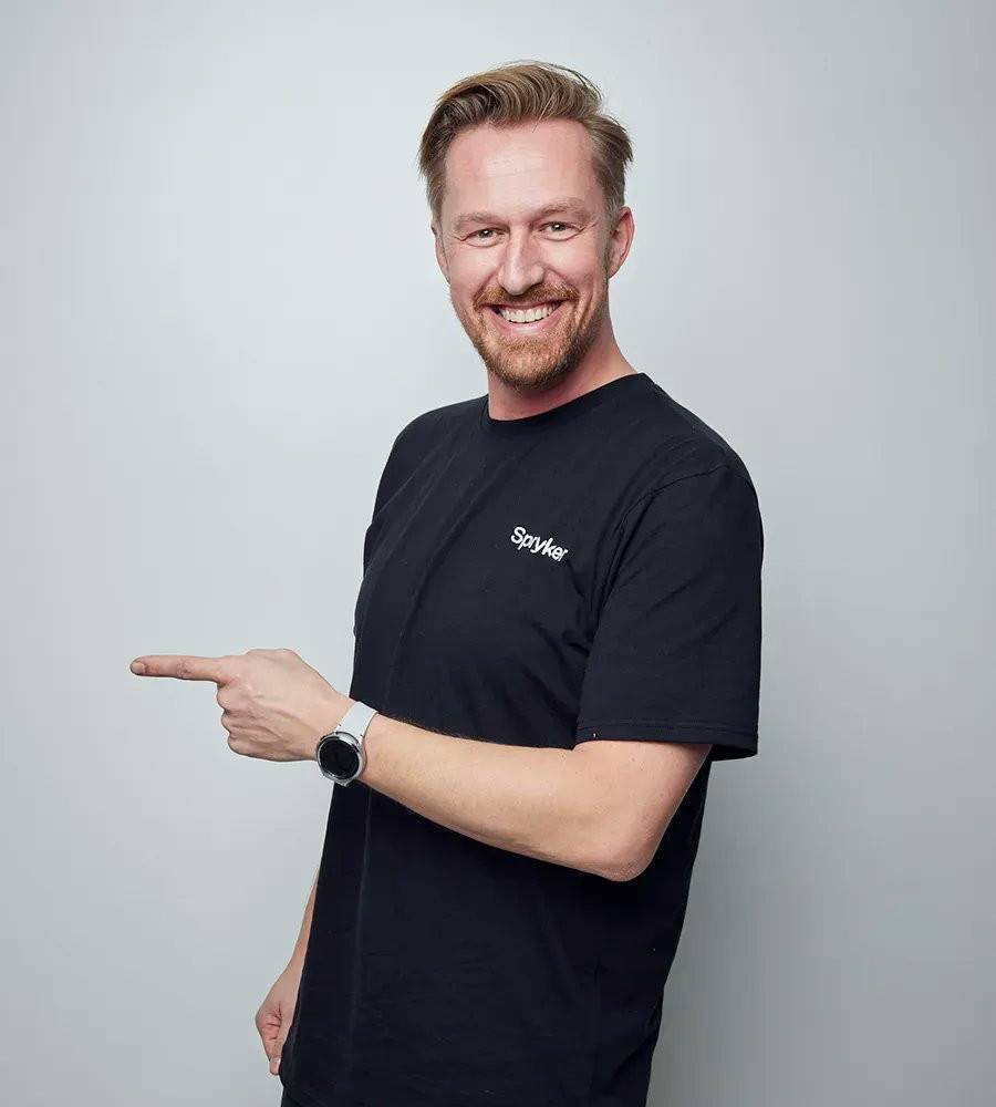A studio portrait of a smiling man in a Spryker t-shirt pointing to the side.