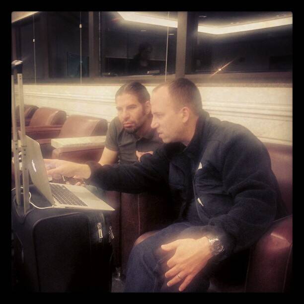 Two men look at a laptop screen while seated in an airport lounge.