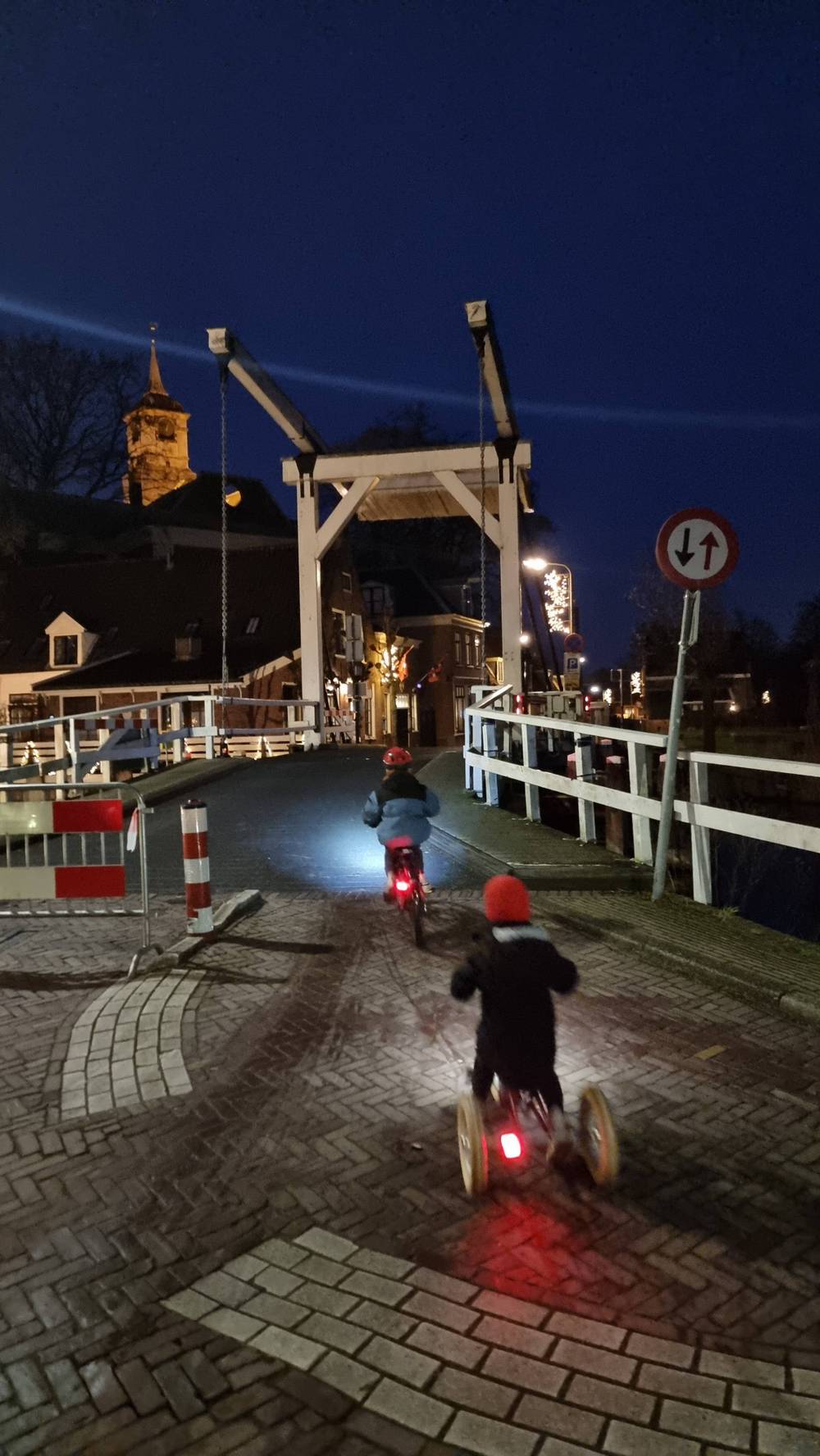 Two children ride bikes with lights across a wooden drawbridge at dusk.