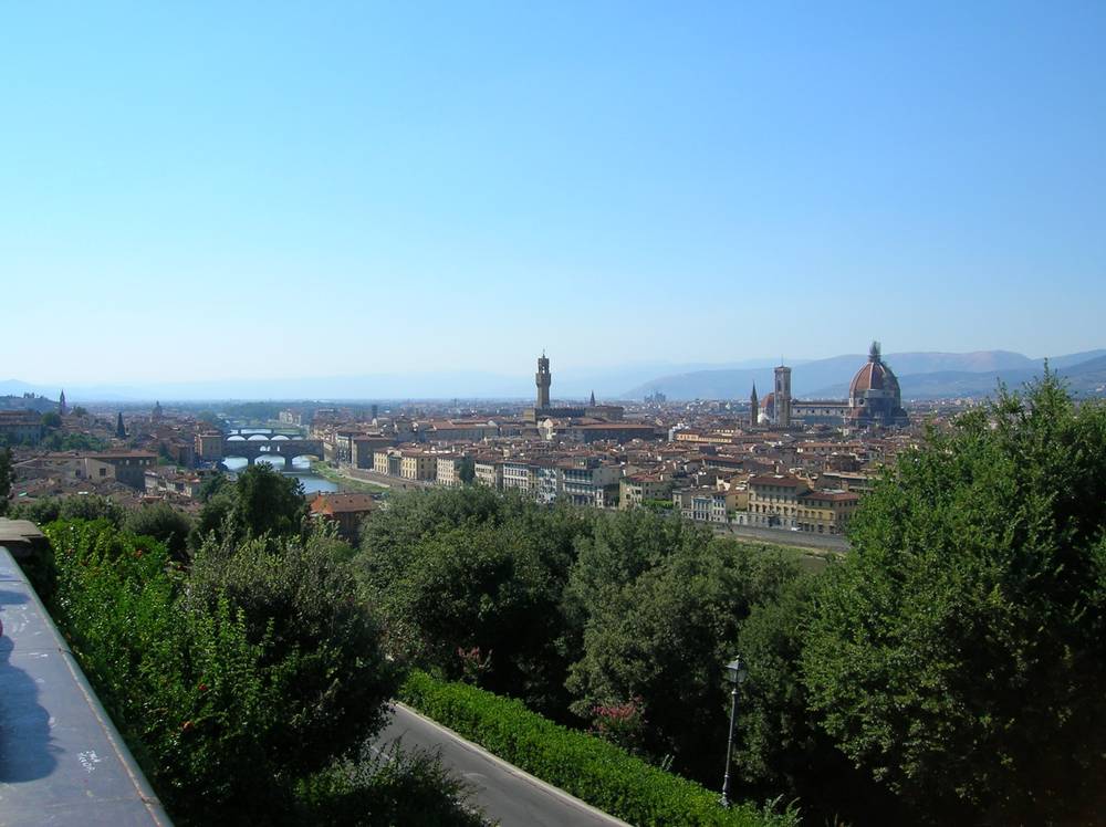 Terracotta rooftops and hills stretch across Florence under a hazy sky, seen from a hillside terrace.