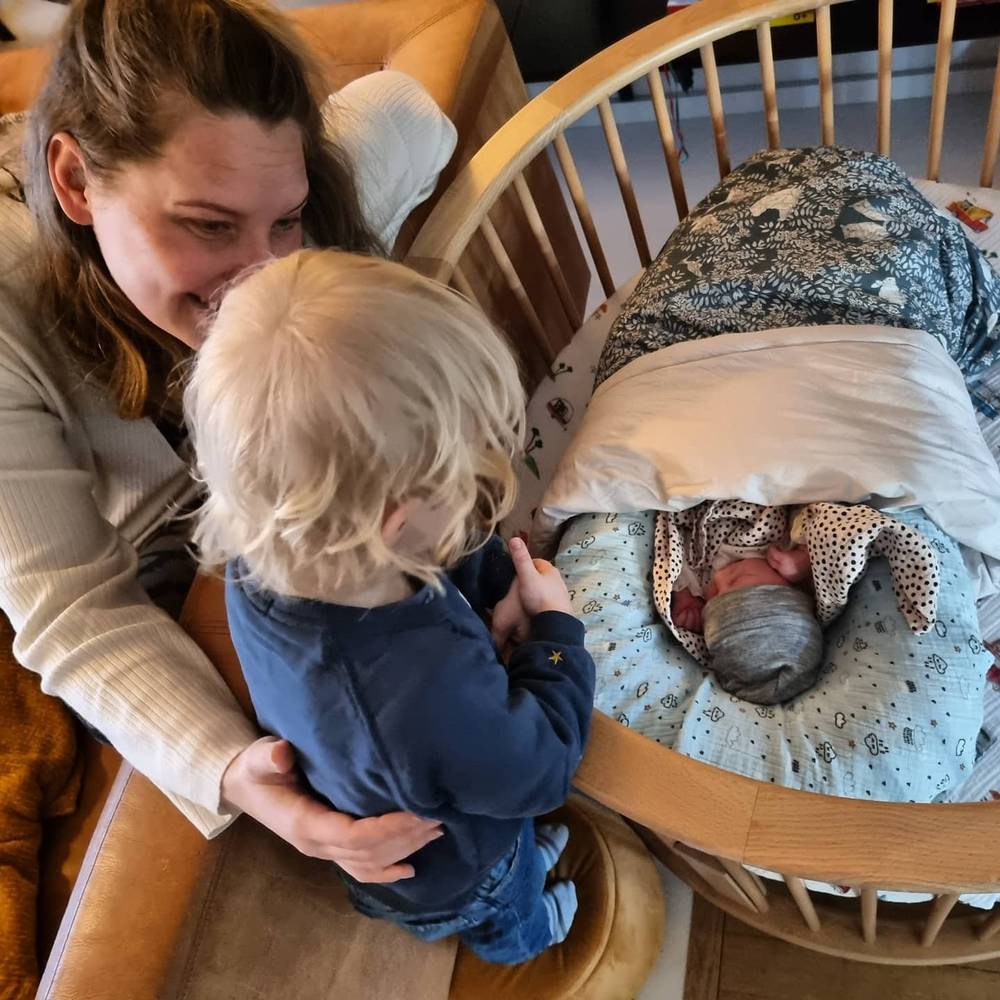 A woman and a toddler both lean over a wicker bassinet to look at a sleeping newborn.