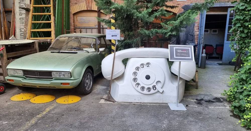 A vintage green car parked beside a giant sculpture of a rotary telephone in a courtyard.